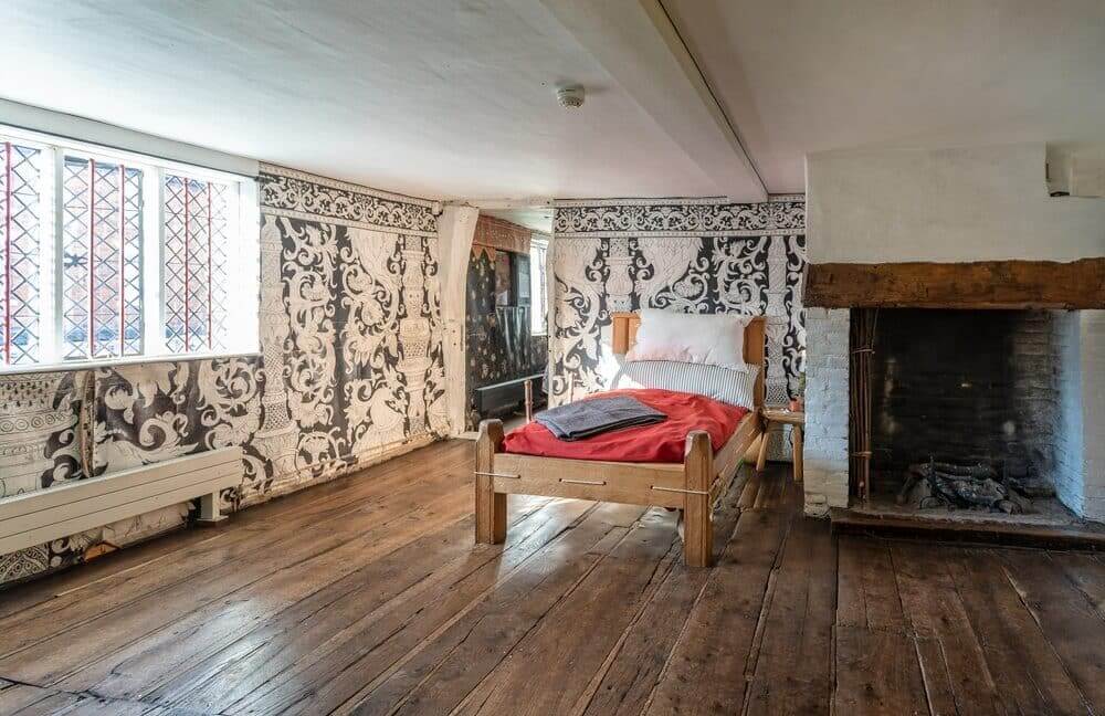 Rustic bedroom with patterned wallpaper, wooden beams, and a large fireplace.