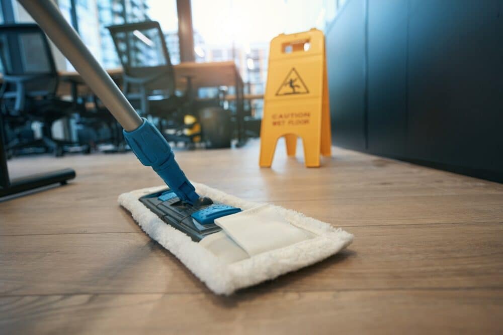 Close-up of a mop cleaning a wooden floor, with a yellow caution sign warning of a wet floor in the background of an office setting.