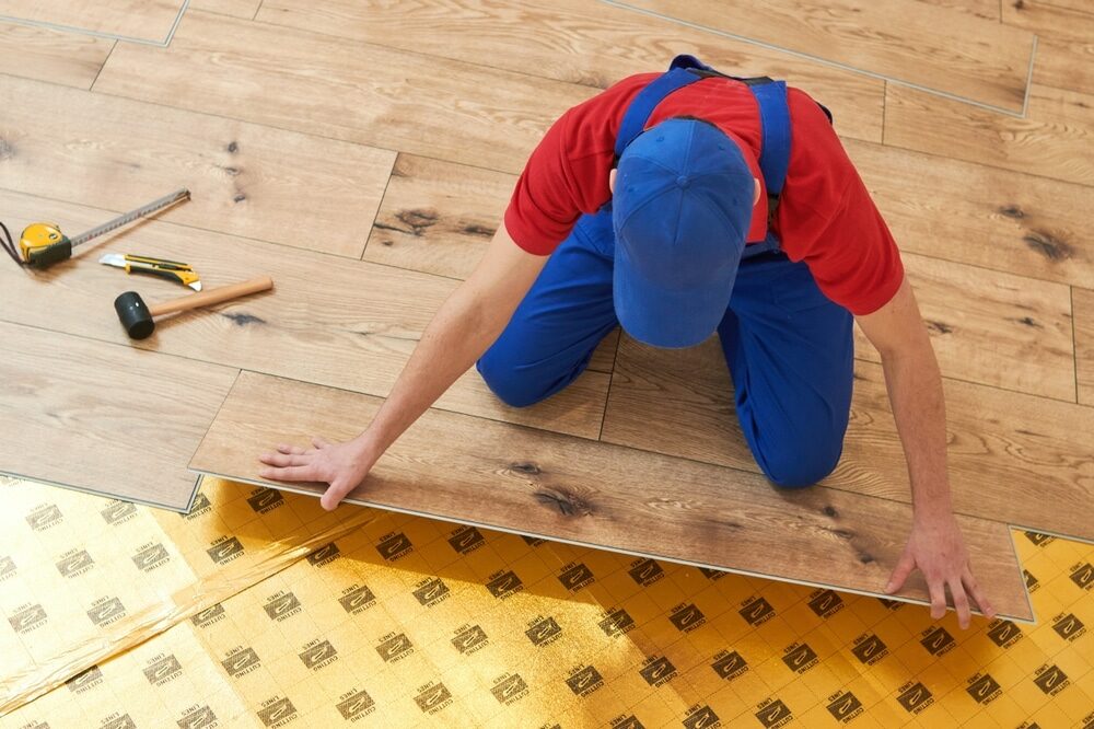 Worker in blue overalls laying laminate flooring over a golden underlayment.