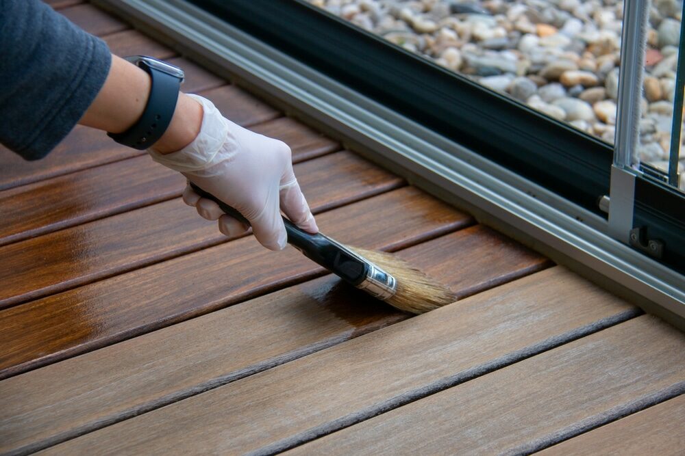 Close-up of a hand in a latex glove applying stain to wooden decking with a brush.