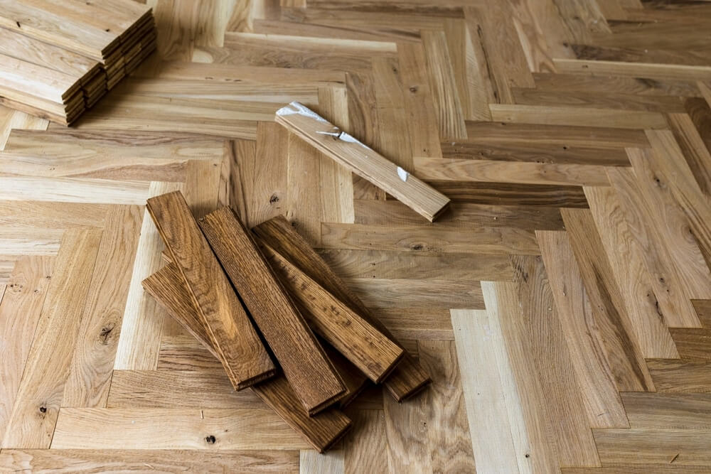 A partially installed herringbone-pattern parquet wood floor, with stacks of wooden planks ready for placement. Some planks are light, while others are darker, showcasing the contrast between unfinished and finished wood tones. The installation is still in progress, with tools and materials scattered across the floor.