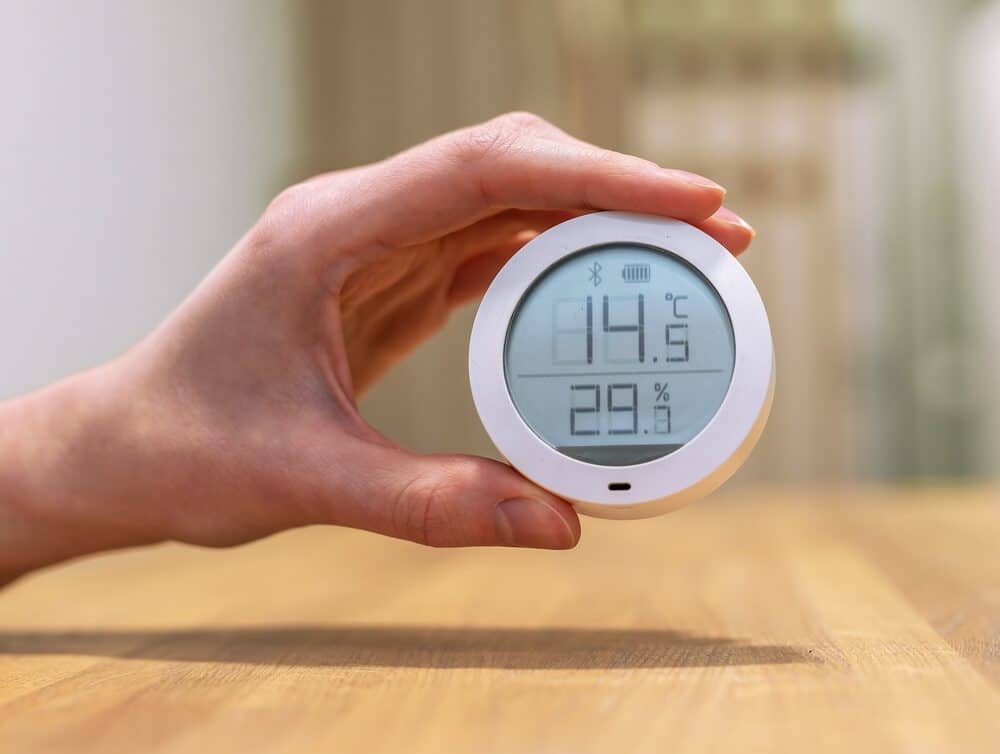 A hand holding a digital hygrometer showing indoor temperature and humidity levels on a wooden table background.