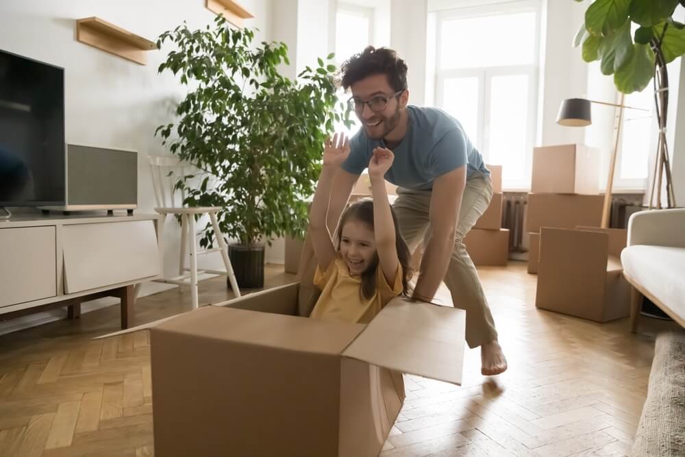 A father plays with his daughter by pushing her in a cardboard box across a beautifully refinished herringbone-patterned hardwood floor. The bright, polished floor enhances the joyful atmosphere in the room, showcasing the expert sanding and refinishing work done by Mr Sander®.