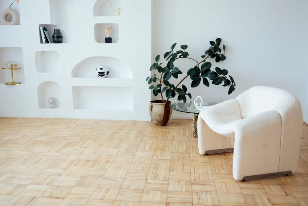 A stylish living room in Islington, UK, featuring a sanded and refinished parquet wood floor by Mr Sander®. The room includes modern decor with a white armchair, a glass table with a plant, and built-in wall shelves displaying decorative items. The parquet floor adds elegance and warmth to the space.