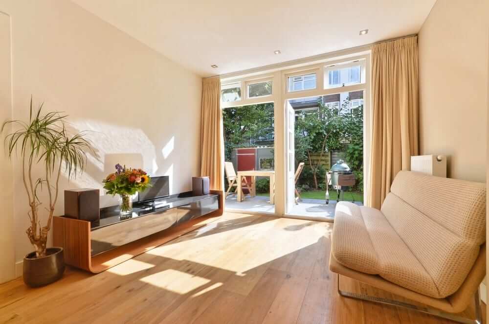 A bright living room with large windows and glass doors leading to a garden. The room features a light oak hardwood floor, a modern entertainment unit, a potted plant, and a comfortable beige sofa.