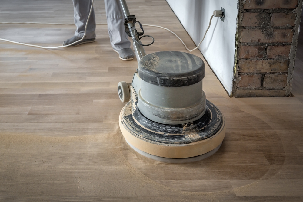 Close-up of a floor polishing machine on a light wooden floor, with a person in the background.