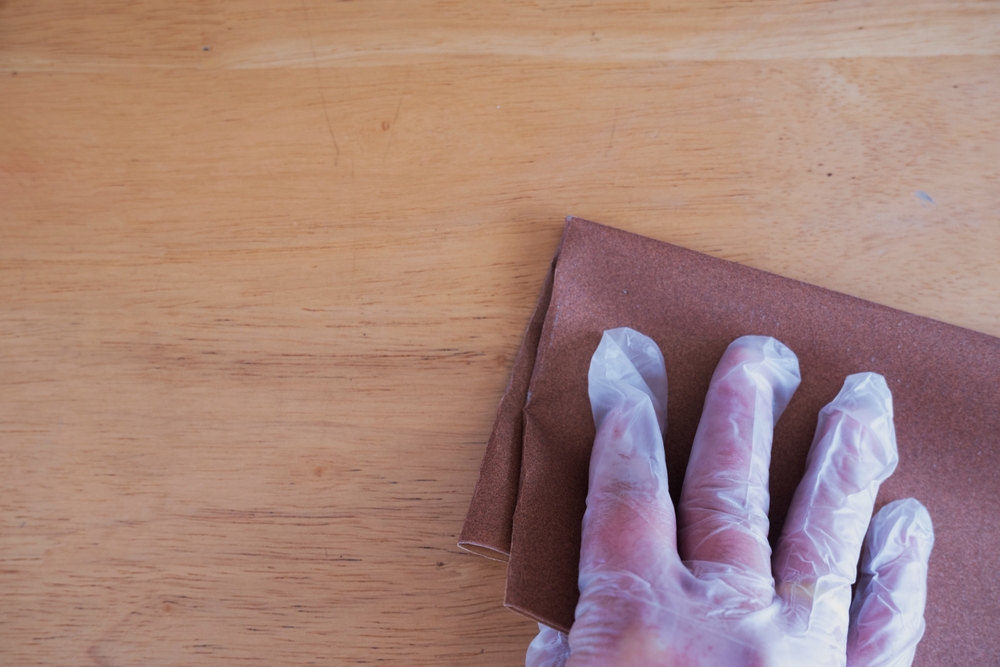 A hand wearing a transparent glove is sanding a wooden surface with a piece of brown sandpaper