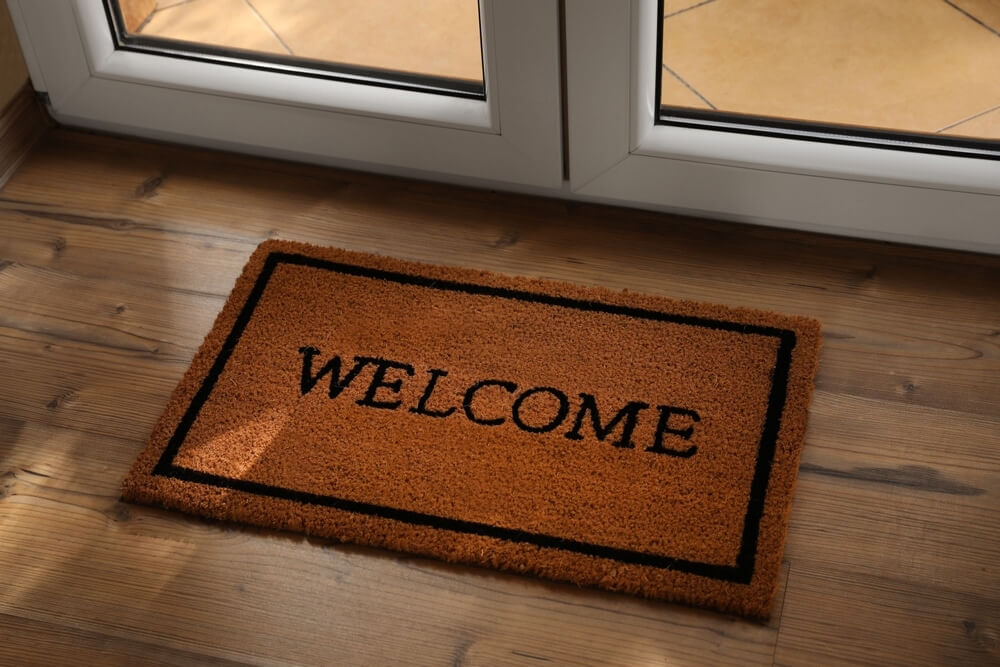 A brown doormat with the word "WELCOME" written in black is placed on a polished wooden floor in front of a glass door. The doormat has a simple design with a black border and serves as a welcoming element at the entrance of the home.