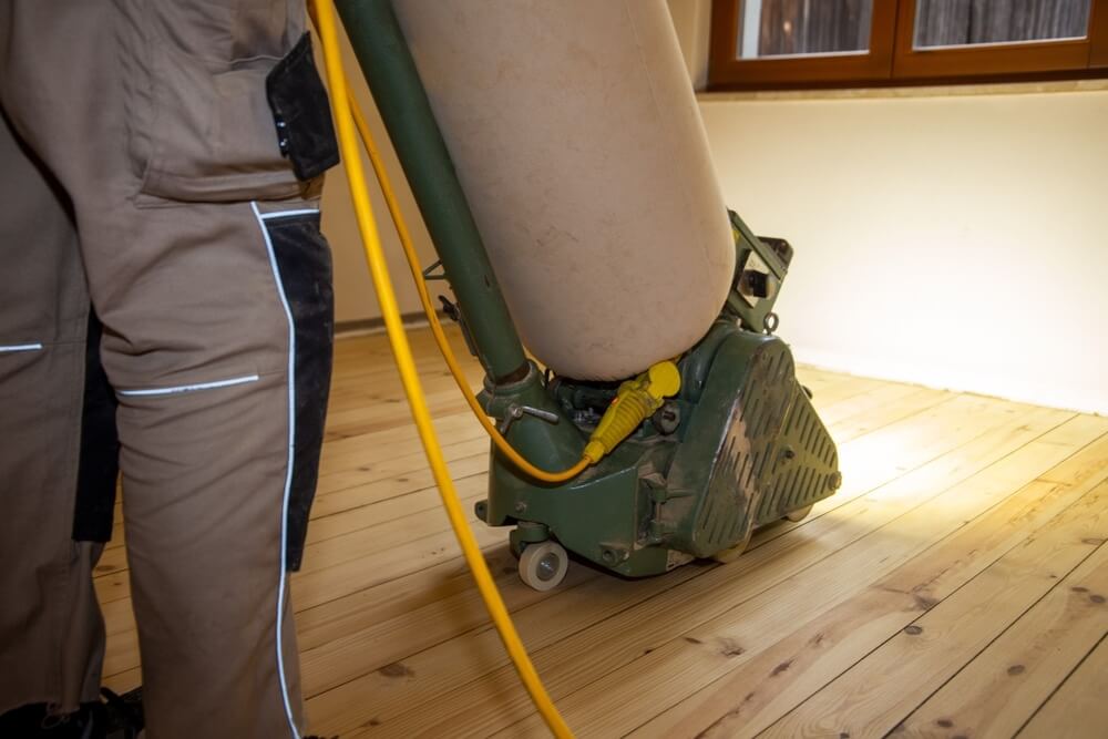 A Mr Sander® technician sands a pine floor in a living room in, UK, using a Lagler HUMMEL® floor sander. The machine features a powerful LED lighting system, high powered aggressive sanding, low dust emissions, quick belt changes, and a 230V AC motor with thermal overload protection.