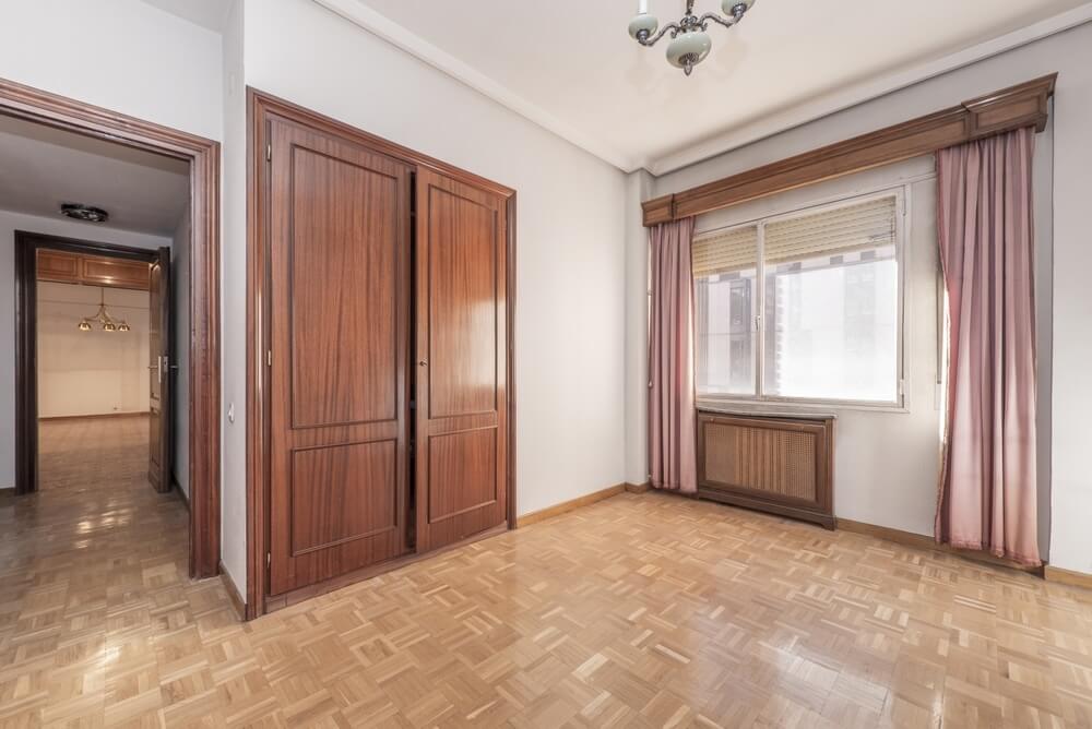 A spacious room in Honor Oak, UK, showcasing beautifully sanded and refinished parquet flooring by Mr Sander®. The room features large wooden wardrobes, pink curtains, and a large window letting in natural light.