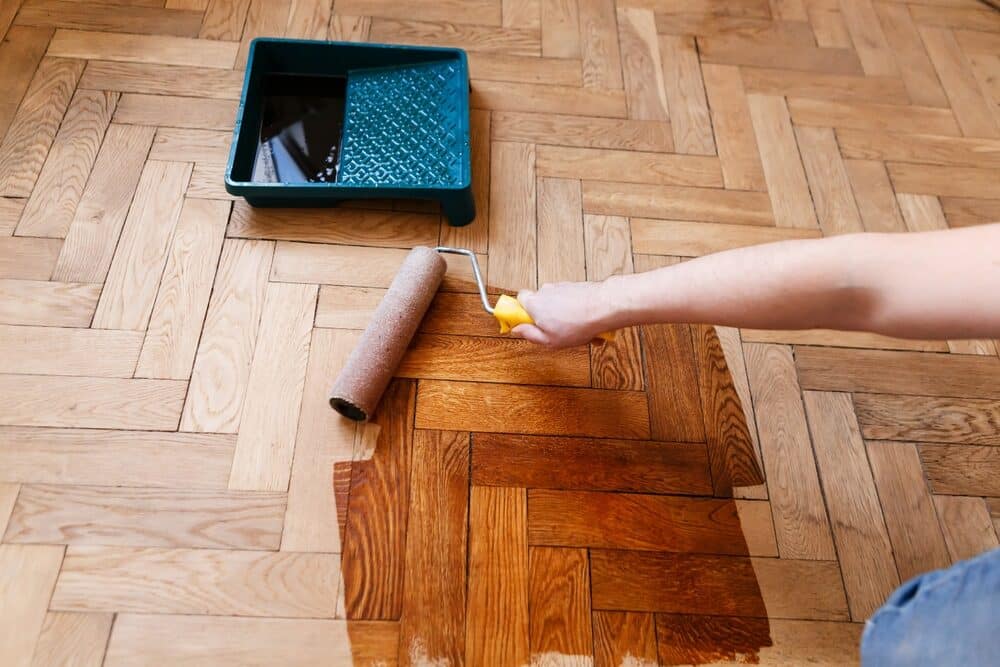 Hand applying varnish on parquet floor with a paint roller and tray.