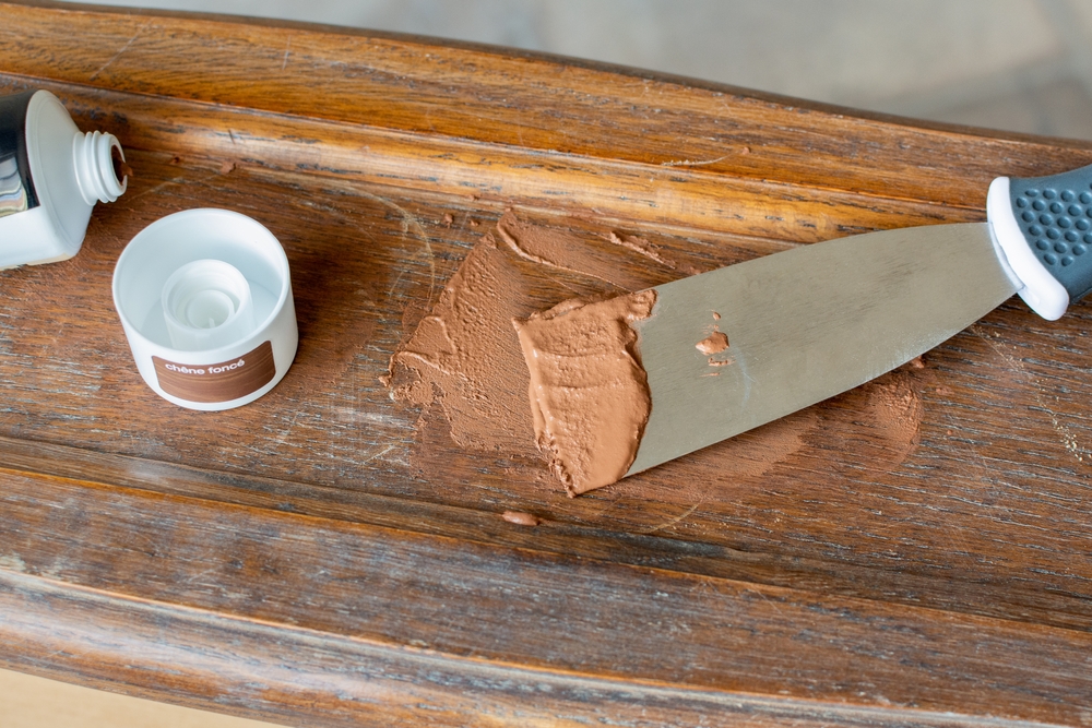A knife spreading wood filler on a wooden surface next to an open container of the filler.