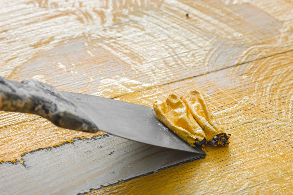 Putty knife scraping off flaky yellow paint from a wooden surface.