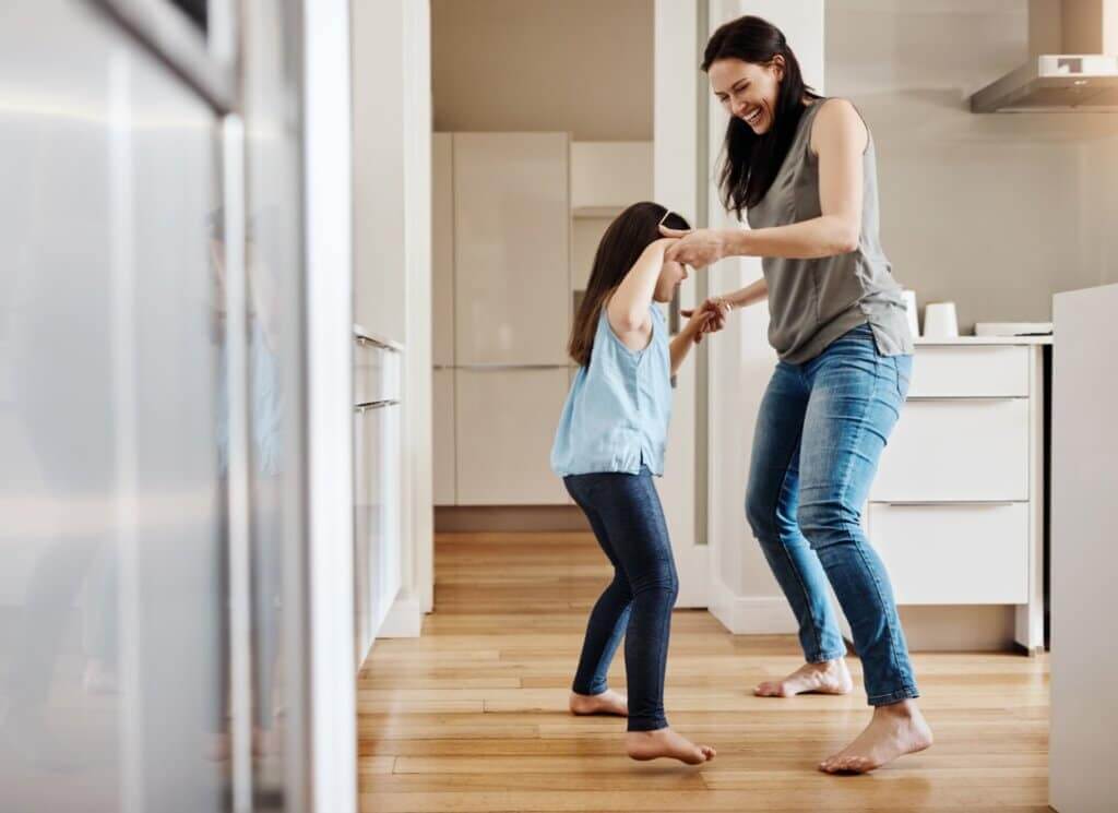A mother and daughter dance barefoot on a beautifully refinished light oak hardwood floor in a modern kitchen. The floor’s smooth and polished surface highlights the high-quality sanding and refinishing work done by Mr Sander®. The room is bright and airy, complementing the warm tones of the wood.