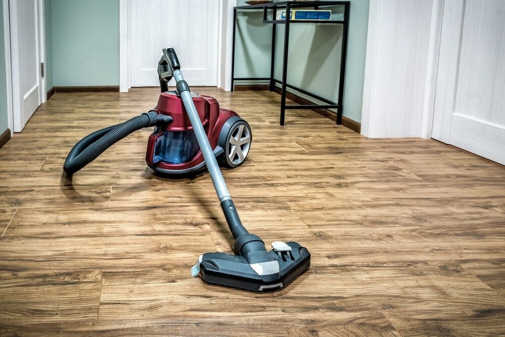 A canister vacuum cleaner with a hose and floor attachment on a wooden floor in a modern home interior.