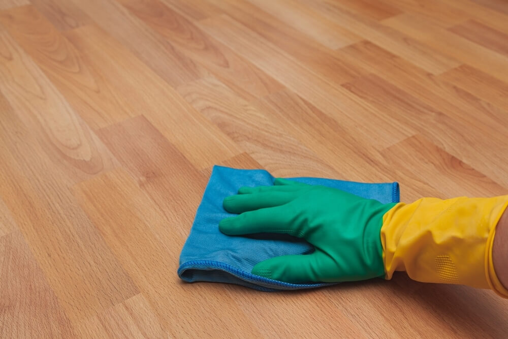 A hand wearing a green and yellow rubber glove wipes a freshly refinished light oak laminate floor with a blue microfiber cloth, ensuring a clean and polished surface after floor sanding and refinishing.