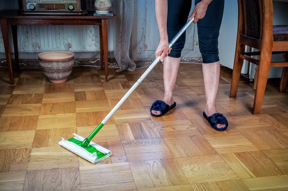 A person is cleaning a light wood parquet floor using a mop with a green handle. The person is wearing blue slippers and casual clothes. The room features wooden furniture, a decorative pot, and an old-fashioned radio on a table, creating a cozy atmosphere. The floor appears smooth and polished, likely after a sanding and refinishing process.