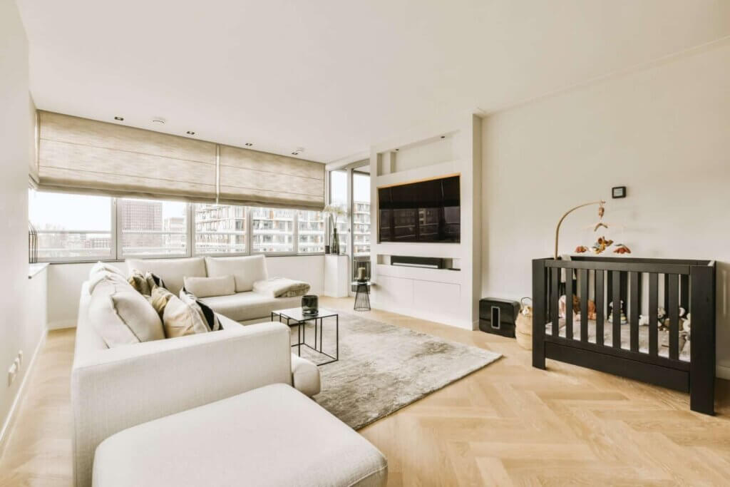 A modern living room with large windows, a white sectional sofa, a flat-screen TV, and a black crib, featuring a newly sanded and refinished herringbone parquet floor. The floor sanding and refinishing were done by Mr Sander® in Beddington, UK.