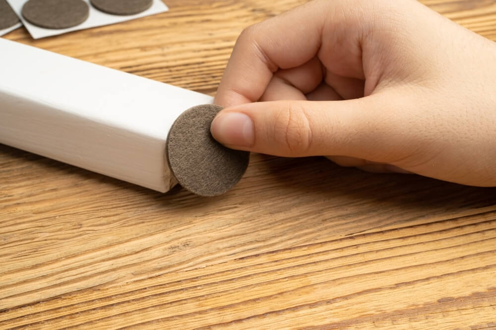 Close-up of a hand applying a felt pad to the bottom of a furniture leg to protect the newly refinished wooden floor. The felt pad provides cushioning and prevents scratches on the floor surface.