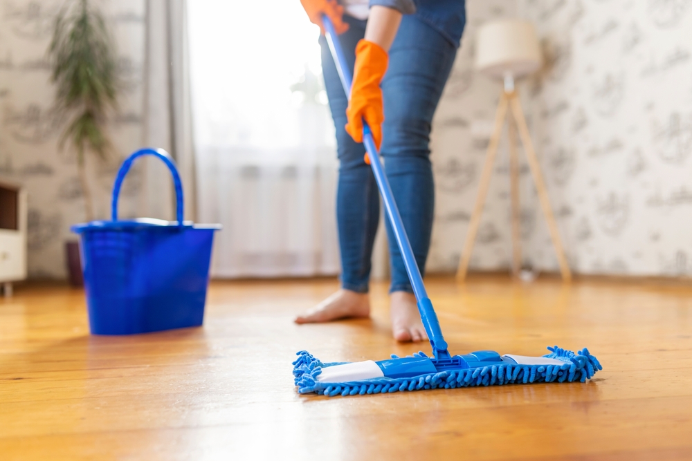 A person in jeans and orange gloves mopping a wooden floor with a blue mop, a blue bucket nearby.