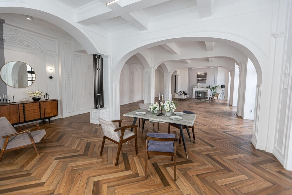 A spacious, elegantly designed room with a beautifully refinished herringbone wood floor. The room features white walls with intricate molding, arched doorways, and coffered ceilings. A modern dining table with flower arrangements and chairs sits in the foreground, while stylish furniture and decorative pieces are placed throughout the space. The polished wood floor enhances the luxurious ambiance of the room.