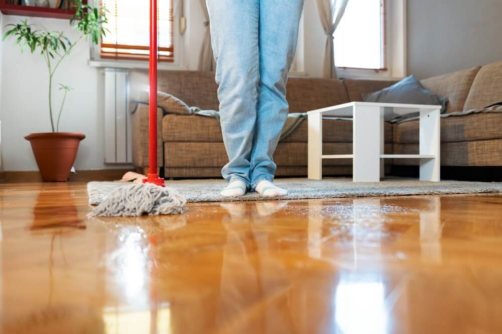 A person wearing jeans and socks is mopping a shiny, recently polished hardwood floor in a cozy living room. The floor reflects the light, highlighting the glossy finish. A sofa, coffee table, and potted plant are visible in the background, adding to the warm, homey atmosphere.