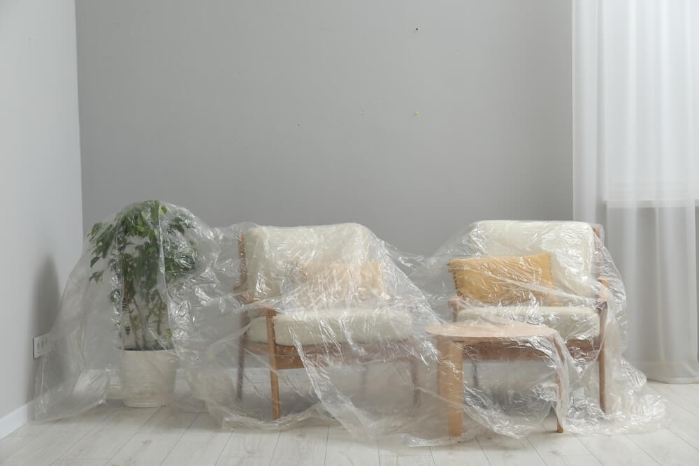 Two armchairs, a side table, and a potted plant are covered with protective plastic in a room with light-colored wood floors. The furniture is prepared for a floor sanding and refinishing project, and the room has a minimalist design with soft natural lighting.
