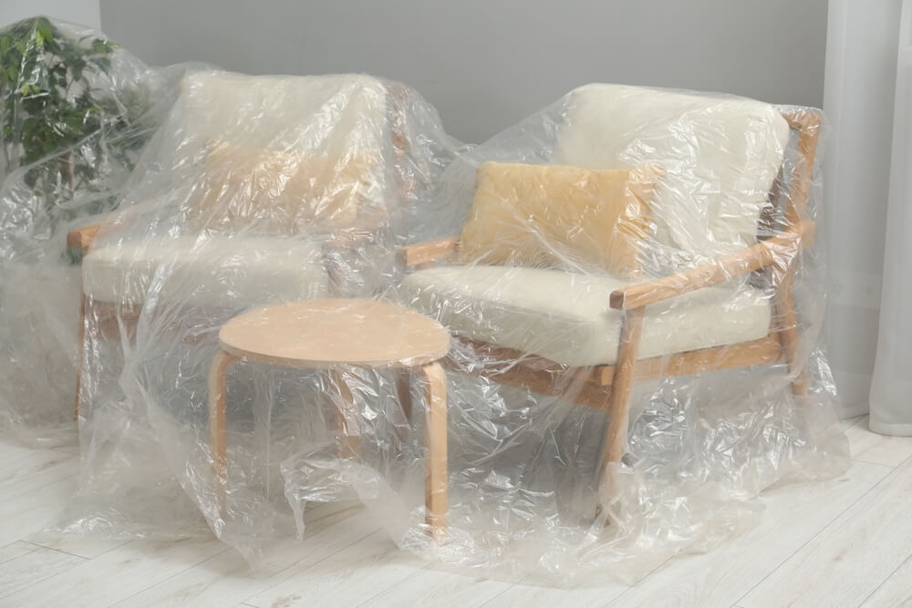  Two cushioned chairs and a small wooden table covered in plastic sheets, protecting them from dust during a floor sanding and refinishing project by Mr Sander®. The room features light-colored, elegant hardwood flooring, showcasing a smooth, polished finish after refinishing. The plastic coverings prevent dust and debris from settling on the furniture while the floor is being professionally restored.
