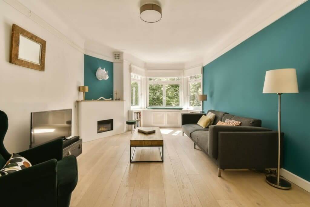 A stylish living room in Tottenham, UK, featuring a freshly sanded and refinished light wood floor by Mr Sander®. The room is tastefully decorated with teal accent walls, modern furniture, and large bay windows letting in natural light.