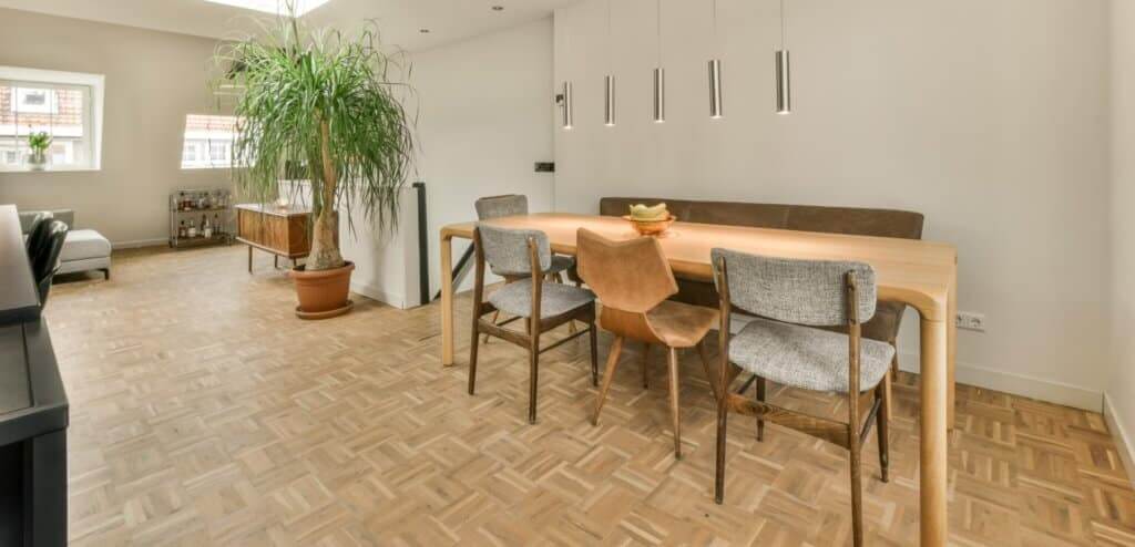  A modern dining area with a beautifully refinished light wood parquet floor. The room is furnished with a wooden dining table, mixed-style chairs, and a potted plant in the corner. Pendant lights hang above the table, and the room has a bright, airy feel with natural light streaming in from the windows. The smooth, polished parquet floor enhances the clean and sophisticated ambiance.