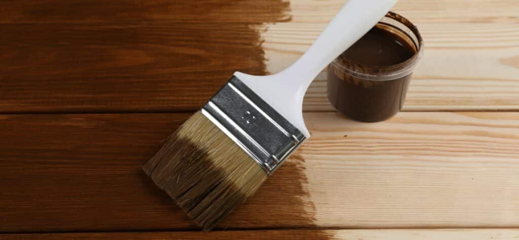A paintbrush with a white handle applying brown stain to a wooden surface, next to an open can of wood stain.