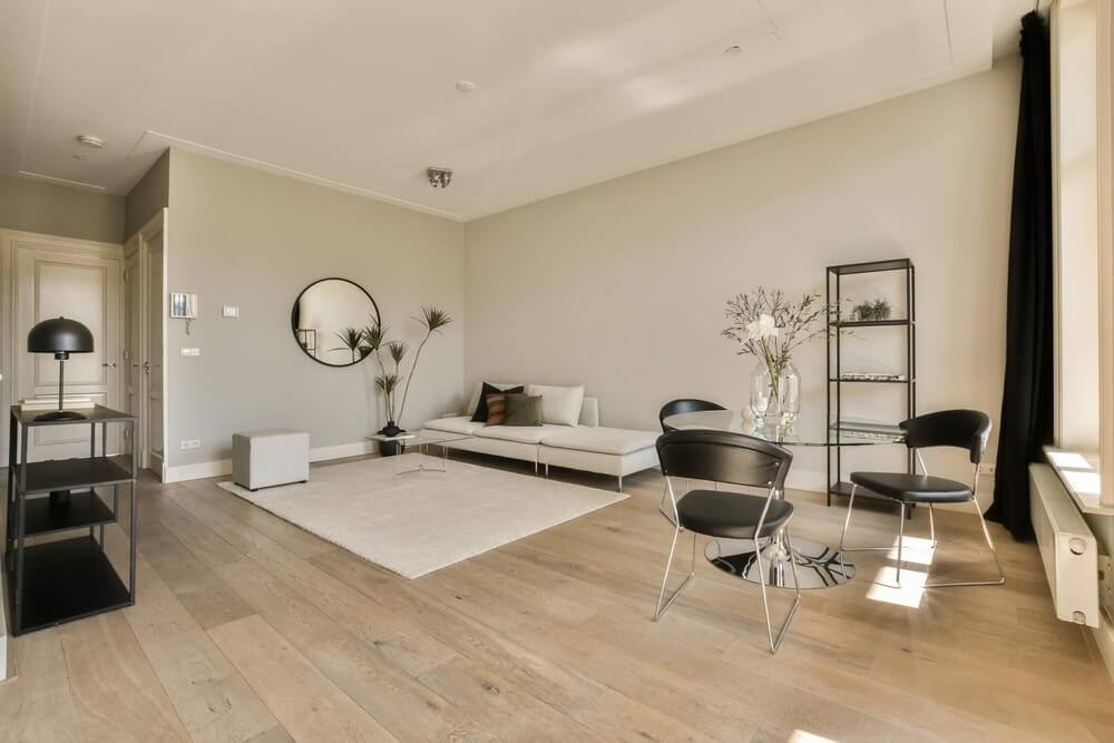Modern living room in Elephant & Castle, UK, featuring light oak hardwood flooring, a white sectional sofa, black chairs, and a glass coffee table. The space is well-lit with natural light and decorated with minimalist furniture and a large mirror. The floor has been expertly sanded and refinished by Mr Sander®, highlighting the natural beauty of the wood.