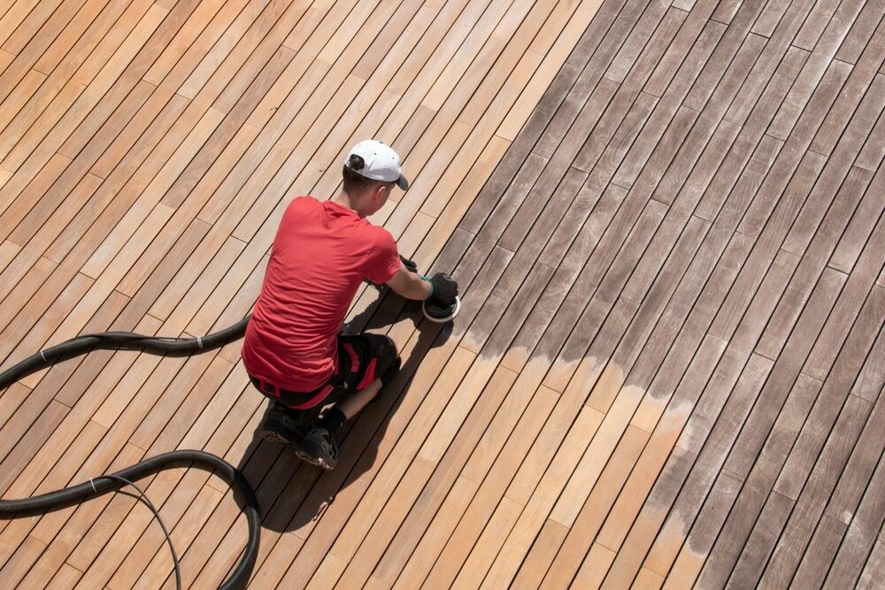 Person in a red shirt using a floor sander on a deck, showing contrast between treated and untreated wood.