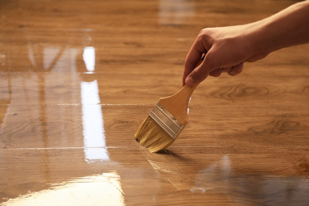 A close-up of a hand applying a clear finish to a wooden floor with a brush. The finish enhances the wood's natural color and grain, giving the floor a glossy and polished appearance.