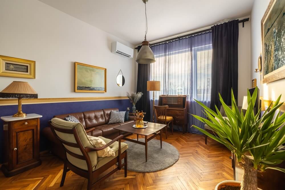 A stylish living room with a mix of modern and vintage decor. The room features a leather sofa, a wooden armchair, a desk, and a large potted plant. The floor is beautifully sanded and refinished in a herringbone pattern.