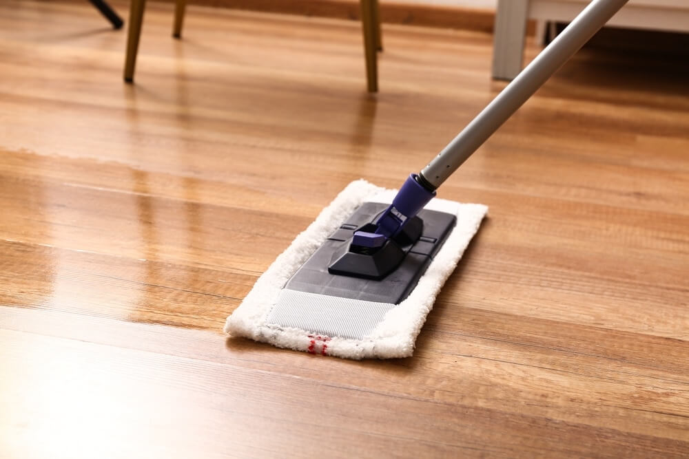 A close-up of a mop being used to clean a polished wooden floor. The floorboards have a smooth, shiny finish, reflecting the light. The mop has a white, fluffy pad attached, indicating gentle cleaning on the well-maintained hardwood surface.