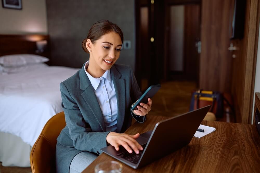 A woman in a business suit is seated at a desk in a hotel room, working on a laptop and holding a phone. The room features a bed in the background and wooden furniture, including a polished wooden floor with a rich, smooth finish that complements the modern decor.