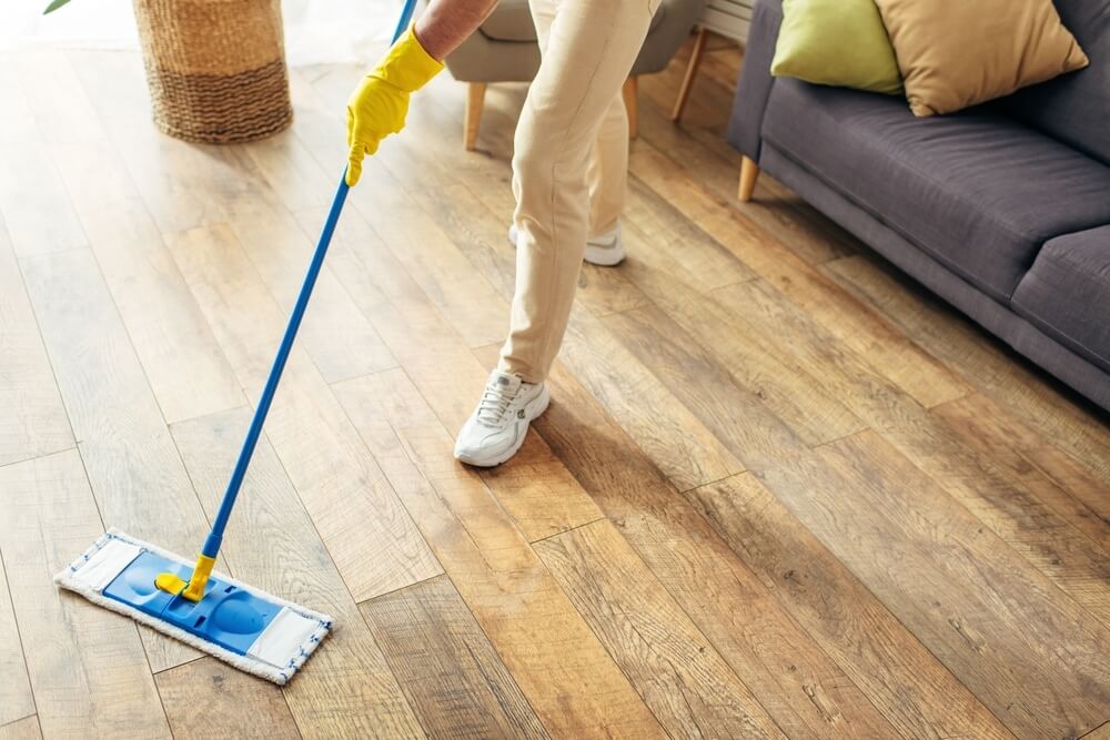 A person wearing yellow gloves is seen mopping a wooden floor, which has been sanded and refinished by Mr Sander®. The individual is holding a blue and yellow mop, and only their lower body is visible. The room features a gray sofa with green and beige cushions, and a wicker basket in the background.