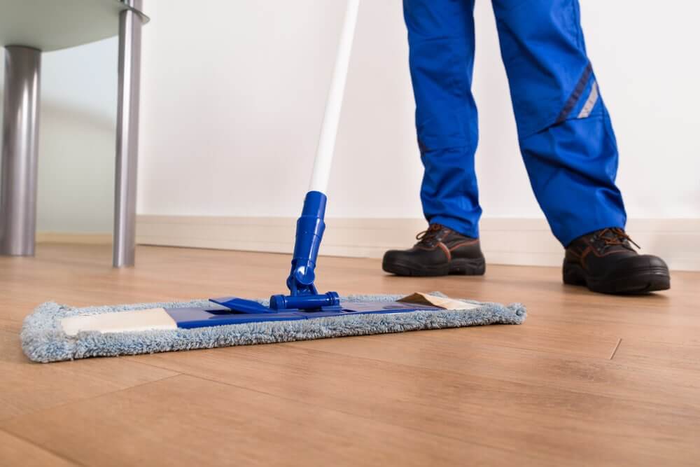 A professional from Mr Sander® mopping a newly refinished wooden floor. The person is wearing blue work pants and black safety shoes, and the mop has a blue handle and a microfiber head, ensuring the floor remains clean and polished.