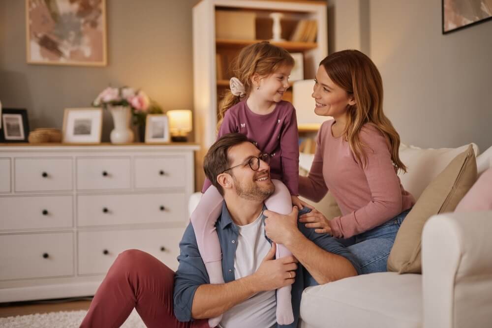 A happy family enjoying time together in a cozy living room. A father sits on the floor with his daughter on his shoulders, while the mother leans on the sofa, smiling at them. The room features soft lighting, a white dresser, and a light-colored wooden floor.