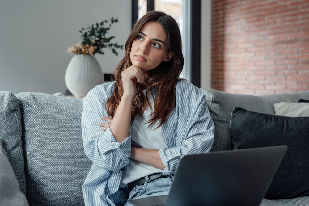 A thoughtful woman sits on a gray sofa with a laptop on her lap, looking contemplatively into the distance. The modern living room features a mix of textures, including a brick accent wall and a white vase with greenery in the background. The muted tones of the décor complement her casual outfit of a striped shirt and jeans, creating a cozy and serene atmosphere.