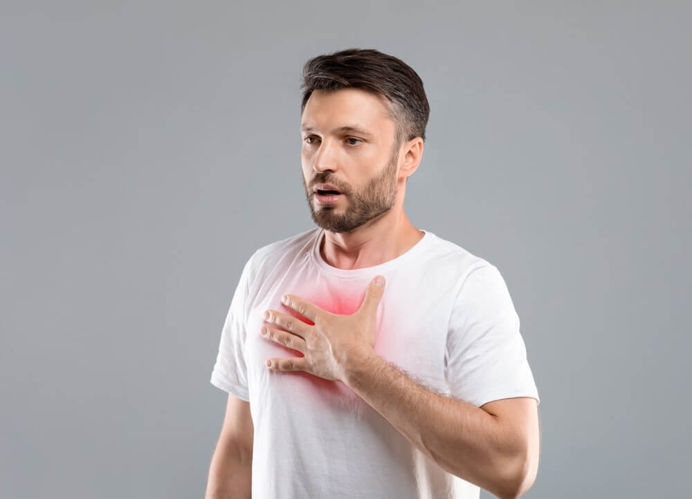 A man in a white shirt holds his chest in discomfort, showing symptoms of potential respiratory issues such as shortness of breath, which could be caused by dust inhalation during floor sanding.