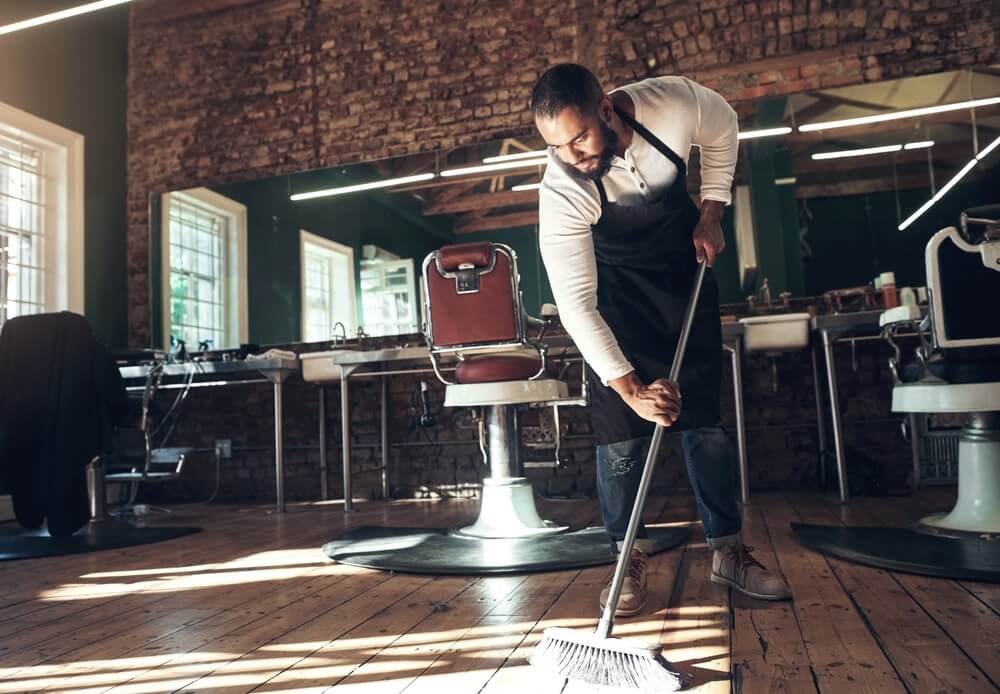 A barber sweeping the floor of a stylish barbershop with exposed brick walls, vintage barber chairs, and large mirrors. The wooden floorboards are polished and well-maintained, reflecting the light from overhead fixtures, adding to the rustic and modern vibe of the space.