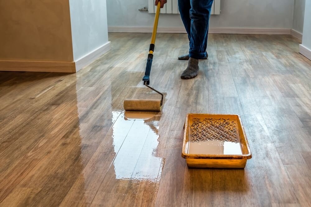 A person using a paint roller to apply varnish to a hardwood floor, with a tray of finishing solution visible in the foreground.