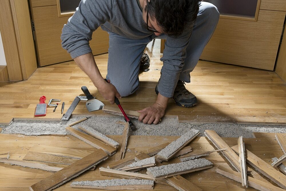 A skilled worker wearing protective gloves is meticulously repairing a damaged hardwood floor using a powerful sanding machine. The machine's abrasive sanding pad is expertly smoothing out the surface of the floor, removing scratches, dents, and other imperfections to restore its pristine condition. This careful restoration process prepares the hardwood floor for refinishing, enhancing its natural beauty and durability.