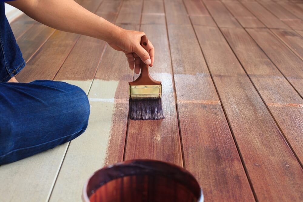  A person is sitting on the floor, applying a dark wood stain to wooden planks using a wide brush. The stain is being applied in smooth, even strokes, and the difference between the stained and unstained sections is clearly visible. A container with more stain is in the foreground.