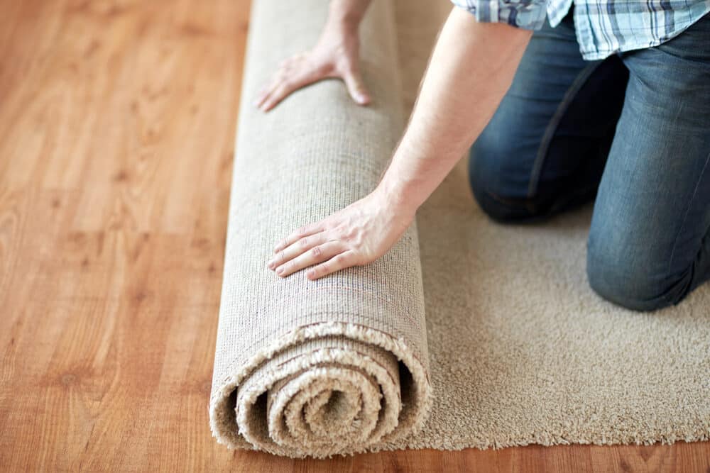 A person unrolling a beige carpet over a wooden floor.