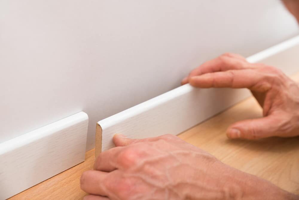 Close-up of hands installing a white baseboard on a laminate floor.