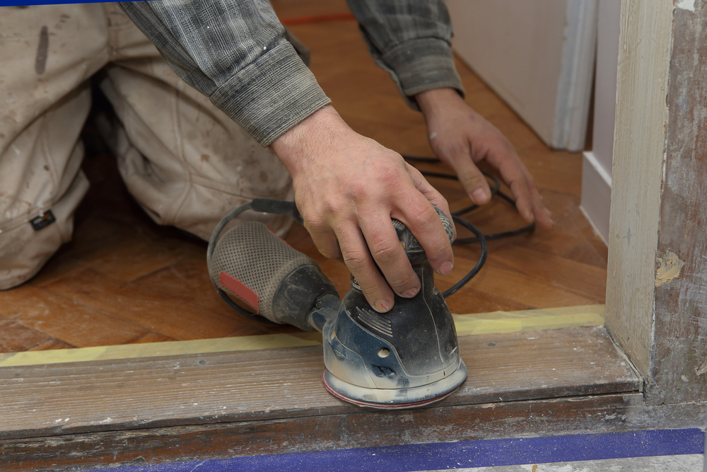 Close-up of a person’s hands using a handheld electric sander on a wooden floor edge.
