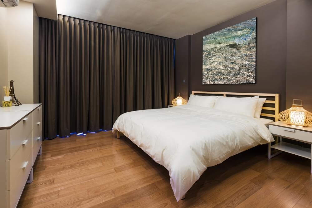 A stylish bedroom showcasing a beautifully refinished hardwood floor, adding warmth to the space. The room features a neatly made bed with white bedding, decorative pillows, and a piece of art on the wall. Soft, dark curtains frame the window, and the wooden floor complements the modern aesthetic. This inviting atmosphere highlights the quality craftsmanship in floor sanding and refinishing completed by Mr Sander® in Buriton, UK.